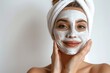 © Marina Demidiuk - Young woman applying cream facial mask to her face gently. White bath towel on a head. Close-up. Girl is caring herself, touching cheek. Light background. Spa, body care, skincare, beauty aesthetic.