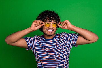 Wall Mural - Photo of young mexican cool curly haired funny man trying new stylish sunglass student in striped t shirt isolated on green color background