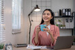 © amnaj - Young woman enjoying coffee at her desk, working from home with a laptop and documents, looking content and productive in a cozy home office setup.