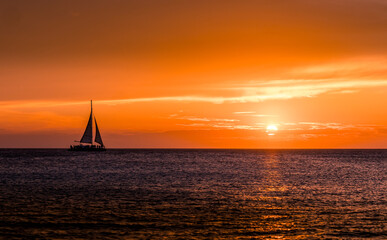 Stunning sunset at Eagle Beach, Aruba, with a sailboat silhouetted against the vibrant orange sky and the serene ocean horizon. A picturesque moment capturing the beauty of the Caribbean.