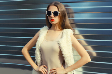 Wall Mural - Modern stylish young woman in white fur vest, glasses, fashionable girl posing on gray background