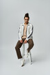 © LIGHTFIELD STUDIOS - A young African American man sits casually on a white stool, wearing a white jacket and brown pants, posing against a grey background.