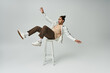 © LIGHTFIELD STUDIOS - A young African American man in a stylish outfit poses dramatically on a grey background, sitting on a stool with one leg extended.