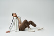 © LIGHTFIELD STUDIOS - A young African American man in a stylish white jacket, brown pants, and white sneakers poses casually on a grey background.