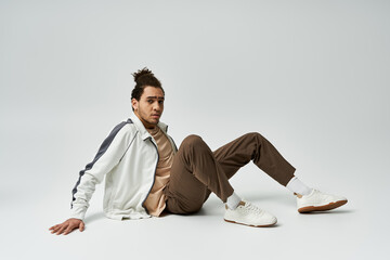 Wall Mural - Stylish young African American man in white jacket with grey stripes, brown pants, and white sneakers poses on grey background.