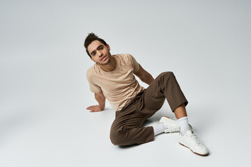 Wall Mural - A young African American man sits on a grey background wearing a beige t-shirt and brown pants.