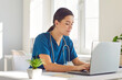 © Studio Romantic - Portrait of a young female doctor or nurse in blue medical uniform sitting at the desk and working in medical office. Woman physician wearing stethoscope in clinic using laptop and typing.