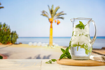 Sticker - Jug of still mineral water with ice and fresh mint   on wooden board table and rocky seaside and tropical beach background.