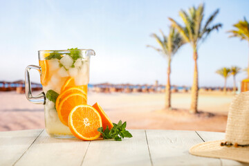 Sticker - Glass of mocktail drink with ice and citrus fruit on a wooden table with sandy tropical beach and blue sky background. Summer table and copy space for products and food and drinks and fruit.