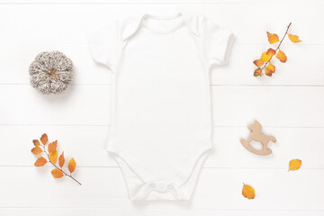 Autumn mockup of white baby bodysuit on wood background with yellow leaves, seasonal decor and toys, flat lay. Blank baby clothes template.