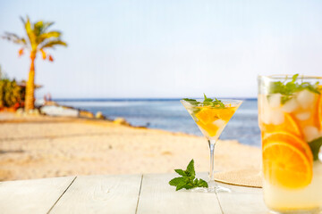 Sticker - Glass of mocktail drink with ice and citrus fruit on a wooden table with sandy tropical beach and blue sky background. Summer table and copy space for products and food and drinks and fruit.