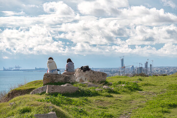 Two young women contemplating cityscape view from Amathus ruins on green hill. Limassol, Cyprus