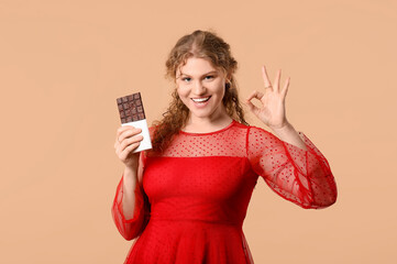 Wall Mural - Beautiful young woman with sweet chocolate bar showing ok gesture on brown background