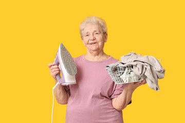 Wall Mural - Happy senior woman with iron and basket of laundry on yellow background
