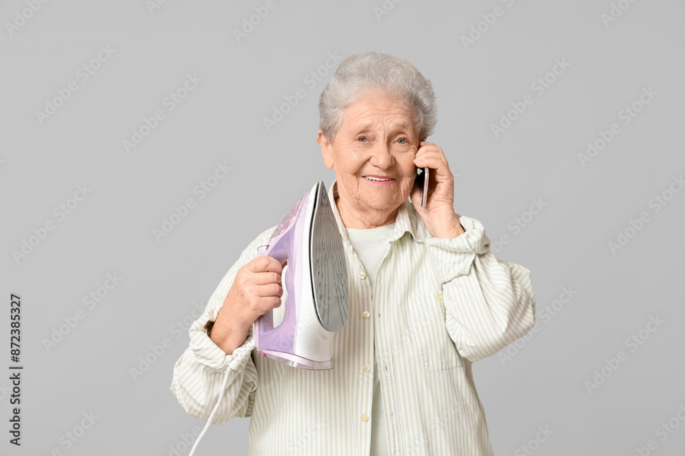 Happy senior woman with iron talking by mobile phone on grey background