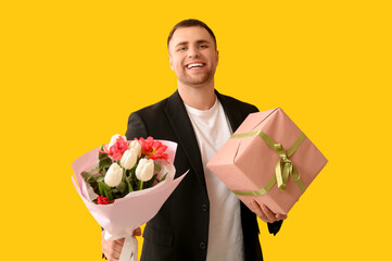 Wall Mural - Handsome young happy man with gift box and bouquet of tulips on yellow background