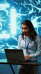 Poster - Vertical video Technician in high tech workspace managing call center tasks, speaking into headset while doing monitoring with laptop. AI company employee talking with clients through headphones mic