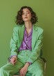 © NiK0StudeO - A fashionable beautiful confident woman wearing a green suit and purple shirt, sitting on a chair with legs crossed against a green background. studio shot captures a minimalist and modern aesthetic