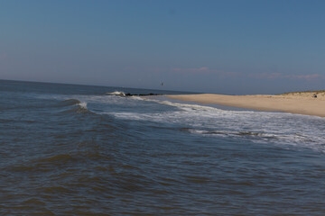 Wall Mural - Beautiful image of the Jersey shore. The white froth of the waves crashing in to the beautiful brown sand of the beach. A small jetty with black rocks can be seen. The blue sky has some white clouds.