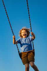 Kid swinging on chain swing on city kids playground. Swing ride. Cute child having fun on a swing on summer sky background. Kids playing in park. Blonde little boy swings at kid playground.