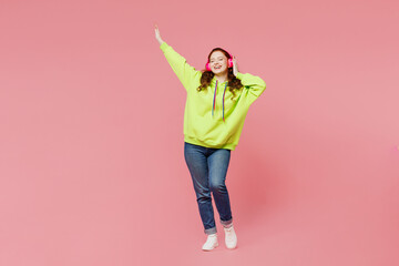 Wall Mural - Full body young smiling happy cheerful ginger woman she wear green hoody casual clothes listen to music in headphones isolated on plain pastel light pink background studio portrait. Lifestyle concept.