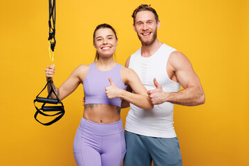 Wall Mural - Young happy strong fitness trainer instructor sporty two man woman wear blue clothes spend time in home gym use trx loops show thumb up isolated on plain yellow background. Workout sport fit concept.