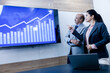 © GAJENDRRA BHATI  - young indian professional woman with businessman discussing trading and stocks in office. Stock brokers looking at indexes graphs on big computer screen