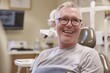 © Olsek - Happy senior man smiling in a dental clinic, ready for check-up. Image shows an uplifting moment. Perfect for promoting oral healthcare, dental practices and senior wellness. Generative AI