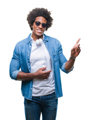 Poster - Afro american man wearing headphones listening to music over isolated background with a big smile on face, pointing with hand and finger to the side looking at the camera.