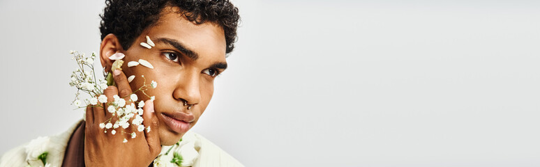 A young African American man poses against a grey backdrop, his face and neck adorned with white flowers, showcasing beauty and diversity.