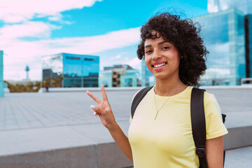 Multiethnic female student wears backpack, does peace victory sign with hand outdoors at city. Young attractive woman with curly hair, broadly smiles. University, education, back to school concept.