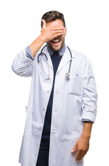 Canvas Print - Young handsome doctor man over isolated background smiling and laughing with hand on face covering eyes for surprise. Blind concept.