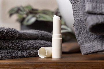 Opened lip balm stick near grey towels, green plant and basin on wood in bathroom, mockup