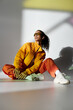 © LIGHTFIELD STUDIOS - A stylish African American woman in a bright yellow jacket and orange pants poses against a gray background.
