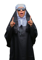 Sticker - Middle age senior catholic nun woman wearing sunglasses over isolated background amazed and surprised looking up and pointing with fingers and raised arms.