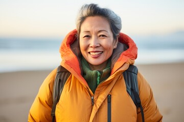 Poster - Portrait of a blissful asian woman in her 60s donning a durable down jacket in front of sandy beach background