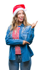 Sticker - Beautiful young blonde woman wearing christmas hat over isolated background with a big smile on face, pointing with hand and finger to the side looking at the camera.