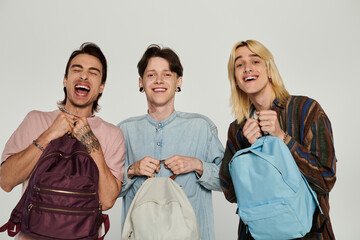 Wall Mural - Three young, queer students in casual attire pose with their backpacks against a gray background.