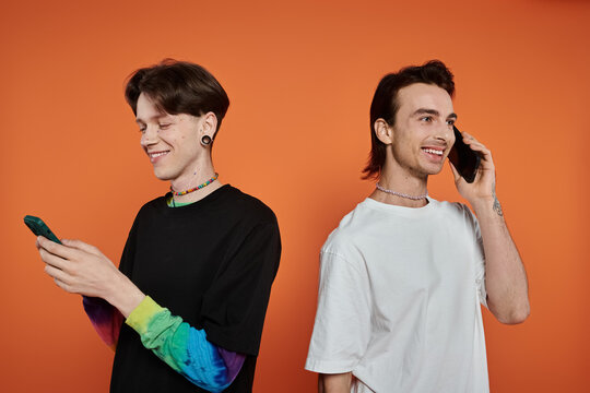 Two stylishly dressed queer friends against a vibrant orange background; one texting while the other is on a call.