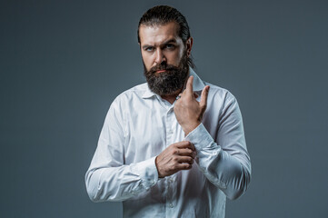 Wall Mural - Man beard putting on a white shirt and cufflinks. Formal wear, putting on cufflinks. Portrait of handsome bearded man in white shirt. Male beard and mustache. Elegant handsome man in white shirt