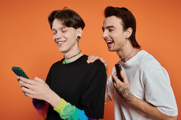 Wall Mural - Two young friends, dressed in vibrant attire, pose against an orange backdrop, sharing a laugh while looking at a smartphone.