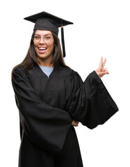 Sticker - Young hispanic woman wearing graduated cap and uniform smiling with happy face winking at the camera doing victory sign. Number two.