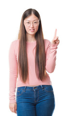 Canvas Print - Young Chinese woman over isolated background wearing glasses showing and pointing up with finger number one while smiling confident and happy.