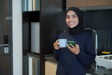 Wall Mural - A woman is smiling and holding a cup of coffee while holding a cell phone. Concept of relaxation and enjoyment, as the woman is taking a break from her daily routine to enjoy a warm beverage