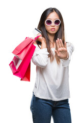 Sticker - Young asian woman holding shopping bags on sales over isolated background with open hand doing stop sign with serious and confident expression, defense gesture