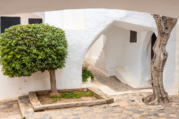 Wall Mural - a narrow stone path between the white walls of the building, at the end of which you can see a illuminated green tree. The atmospheric village of Binibeca on Menorca in Spain