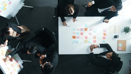 Wall Mural - Top down aerial view of business group sharing and brainstorming idea by using sticky notes at meeting room. Investor writing marketing strategy on board while receive note from manager. Directorate.