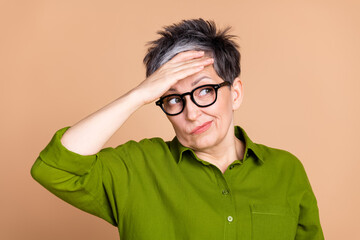 Wall Mural - Photo of lovely pensioner lady look empty space touch forehead confused dressed stylish green garment isolated on beige color background