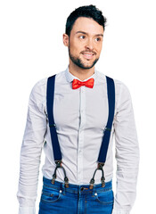 Poster - Hispanic man with beard wearing hipster look with bow tie and suspenders looking away to side with smile on face, natural expression. laughing confident.