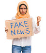 © Krakenimages.com - Young brunette arab woman holding fake news banner annoyed and frustrated shouting with anger, yelling crazy with anger and hand raised